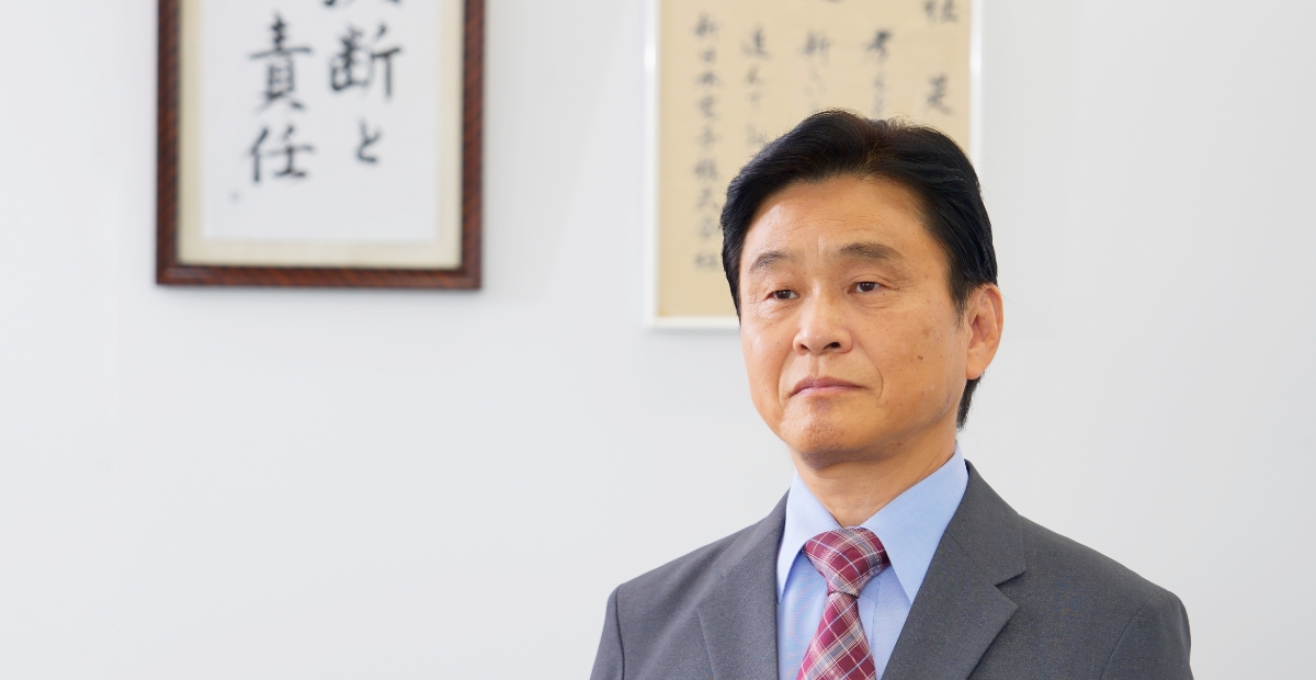 企業理念を背にスーツ姿で佇む日本人経営者