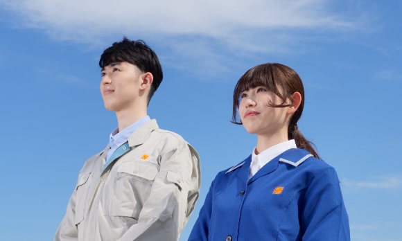 作業服を着た新日本電子の社員が青空の下で前向きに見つめる姿