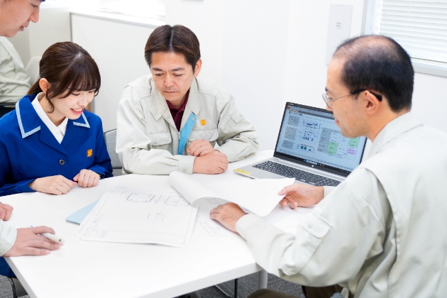 設計図を囲んで打ち合わせをするエンジニアチームと作業服の技術者たち